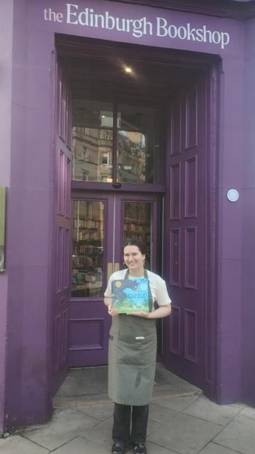 Longshot of bookseller Sarah holding up Julia Donaldson and Sara Ogilvie's picture book, Gozzle, outside the Edinburgh Bookshop