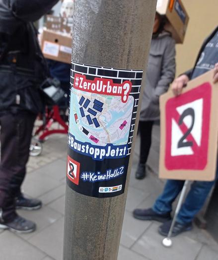 Ein Sticker auf einem Metallpfahl. Auf dem Sticker ist von oben nach unten:
der Hashtag Zero Urban
eine Karte von Ruhrort und einem Teil des Hafens. Darauf eingezeichnet die Hallen 1 und 2, 2 alternative Hallenstandorte mit der Beschriftung NEIN und 8 weitere Hallen über Ruhrort verteilt
der Hashtag Baustopp Jetzt
das Logo der Keine Halle 2, der Hashtag Keine Halle 2, einige Symbole (Fediverse, Mastodon, PeerTube, Prideflag, Extinction)

Im Hintergrund stehen Menschen, die protestieren.