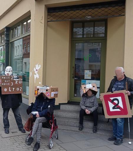 Eine kleine Gruppe von Menschen blockiert teils sitzend, teils stehend den Eingang eines Lokals. Einige tragen bemalte und beklebte Protestkartons und Schilder.