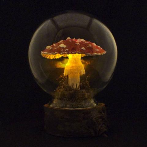 A handcrafted mushroom lamp inside a glass dome. The lamp is shaped like an Amanita muscaria, with a red cap speckled with white spots and a delicate, textured stem. The mushroom emits a warm golden glow from within, illuminating the moss and organic materials at its base. The dome rests on a wooden base with a natural, rustic appearance. The background is dark, making the glowing mushroom the focal point of the image.