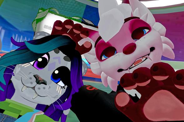 The image shows two anthropomorphic characters posing for a photo.

Left character: Resembles a feline with blue and purple hair, large eyes, and whiskers.

Right character: Resembles a canine with white and pink fur, sharp teeth, and large eyes.

Both characters are raising their paws towards the camera, making a playful gesture. The background is a colorful, indoor setting with various objects and lights, suggesting a fun, playful environment