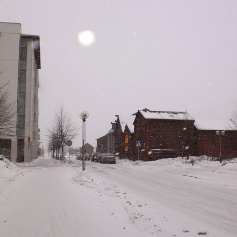 Wintery scene of overcast sky and heavy snowfall in central Oulu, Finland, with some old wooden market square buildings ahead and some modern buildings seen at the left edge.