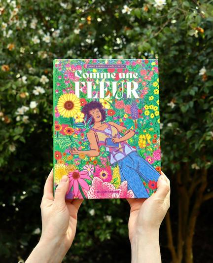 This is an outdoor photo where I’m holding my freshly released graphic novel! I went to the Jardin des Plantes in Nantes to take this photo series and took advantage of the beautiful weather to do so. The background is filled with greenery and flowers. In this first photo, I’m showing the cover of my book.
