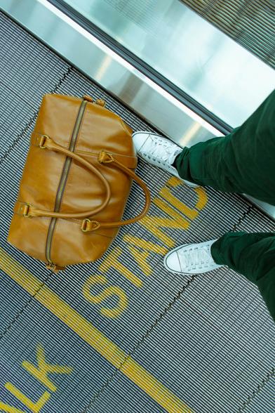 Photograph of a person stranding on a moving walkway in an airport beside a brown leather handbag. The word "stand" is written in yellow at their feet. By White Field Photo, under a free Unsplash license: https://bit.ly/3XNjLyr.