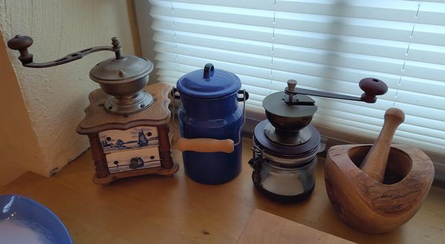 Blick auf ein Holzfensterbrett in der Küche. Darauf, von links nach rechts: eine historische Kaffeemühle, mit blau-weißen Kacheln außen, die Windmühlen und andere Küstenszenen zeigen. Dann eine blaue Milchkanne mit Deckel und Henkel. Dann eine moderne Kaffeemühle mit Unterteil aus Glas. Ganz rechts ein Mörser aus Olivenholz.