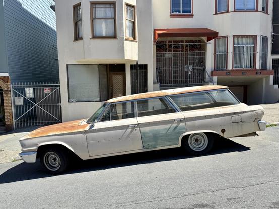 Photograph from a side profile of a vintage station wagon automobile, highlighting its enormously long body, expansive glass windows, and blade-like, creased shaping of its side panels. The car was painted cream and shows extensive patches of rust, sun-bleaching, and other damage to its finish. The car is parked at the curb of a downward-sloping urban street in front of a similarly cream-colored Victorian apartment building.