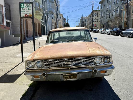 Photograph from a head-on front view of a vintage station wagon automobile, highlighting its expansive hood and wide windshield, four round headlights, and perforated chrome grille. A nameplate spelling #Ford in widely-spaced letters is faintly visible on the front lip of the car’s hood. The vehicle was painted cream but now shows large amounts of rust, sun-bleaching, and other damage to its finish. The car is parked at the curb of a downward-sloping urban street with Victorian-style apartment buildings and other vehicles visible in the sunny background.