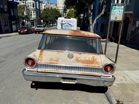 Photograph from a direct rear view of a vintage automobile, highlighting its wide frame, expansive glass rear windshield, pie-like round taillights, and perforated chrome detailing. A decorative badge and nameplate spelling #CountrySedan in streamlined script letters is attached to the station wagon’s rear gate. The car was painted cream but now shows extensive patches of rust, sun-bleaching, and other damage to its finish. The car is parked at the street curb, with trees, houses, and other vehicles visible in the sunny background.