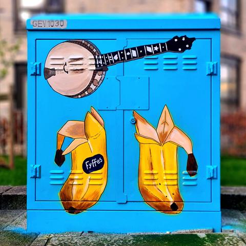 A junction box on a Glasgow street which has been decorated with a painting of a banjo and a pair of banana boots.