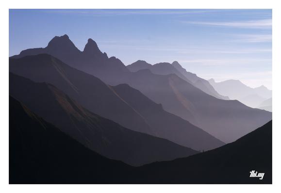 View from a mountain pass into the far distance with ~10 layers of silhouettes of both smooth and craggy mountain ridges, against the hazy late afternoon light in autumn last year. Serene, almost clear sky.