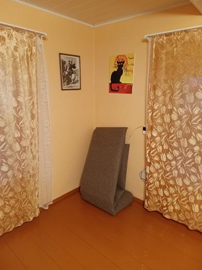 Corner of a room. Two windows hidden by curtains and a wooden floor painted in "floor paint" which is always this colour and only this colour (a fake looking shade of brown). Two pictures hang on the wall, a hand painted cat and some dude on a horse stabbing a lindwurm. There's a new carpet bundled in the otherwise empty corner. Some cables peek out from behind the curtain.