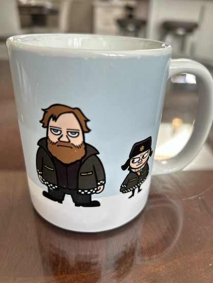 Photo of a mug with the cartoon images of a large bearded man wearing a heavy winter jacket with a black and white grid pattern at the hem line and bottom of the sleeves. He is looking straight ahead in a serious expression. A shorter character is standing to his left wearing the same type of jacket, as well as a winter cap with ear flaps and with what looks like a police star insignia in front. The short character is giving side-eye to the bearded one