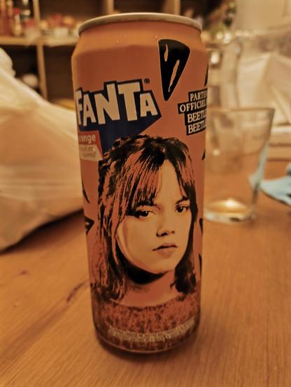 La imagen muestra una lata de refresco Fanta sabor naranja colocada sobre una mesa de madera. En la lata aparece el rostro de una joven con expresión seria, que recuerda al estilo gráfico promocional de la serie Wednesday o Beetlejuice. De hecho, a la derecha del rostro se puede leer “PARTENAIRE OFFICIEL BEETLEJUICE BEETLEJUICE”, lo cual indica una colaboración oficial entre Fanta y la franquicia de Beetlejuice.

La escena parece haber sido tomada en un entorno doméstico: se observan vasos, una bolsa plástica blanca y algunos objetos desenfocados al fondo, probablemente utensilios de cocina o artículos de uso cotidiano. La iluminación es cálida y tenue, lo que da un ambiente acogedor a la imagen.