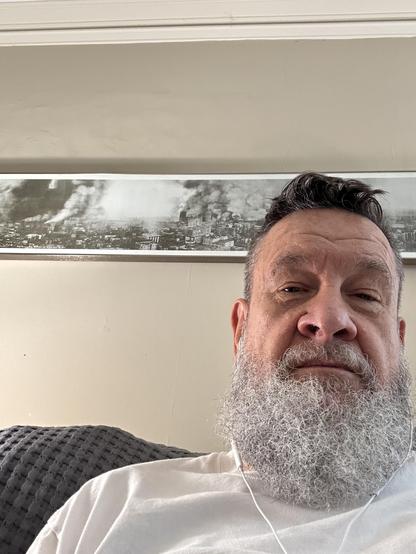 Man (shoulder shot) with short dark hair and long gray beard, in a white tshirt, sitting on a dark gray seat visible slightly on on the left. Behind him is an off white wall with a white crown moldings above. On the wall is a famous panoramic photo of San Francisco (“The Burning City 10am April 18 1906”) after the BIG earthquake…