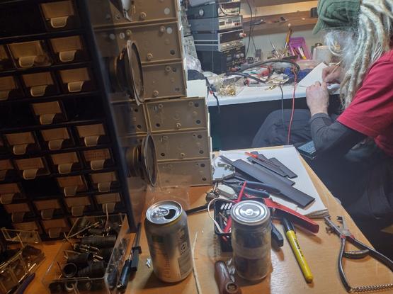 An electronic workshop. Stuff all over. Dude with gray dreads in background
