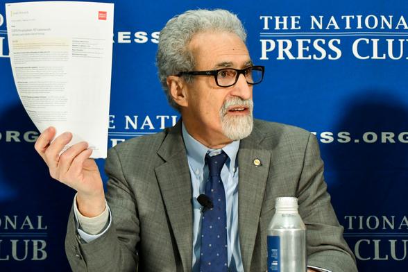 Man in a gray suit, glasses, and goatee holding up a document in one hand.