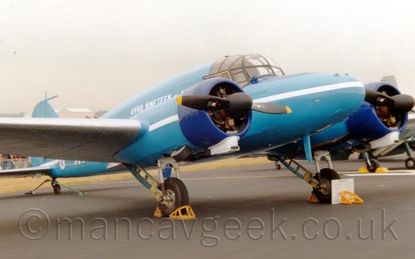 Side view of a blue, twin propellor-engined, World War 2-era transport aircraft parked facing to the right.
There is a white stripe running along the body and the wing-mounted engines, with white "Avro Nineteen" titles on the upper fuselage.
There are yellow metal chocks in front of and behind the main wheels, with a blue metal support around the tail-wheel.