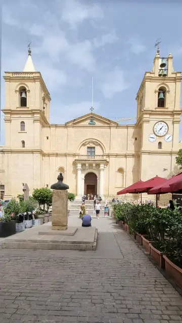 Die Kathedrale "Saint Johns Co-Cathedral" von frontal fotografiert – eine sandsteinfarbige große Kirche mit drei Uhren, die die Uhrzeit, Wochentag und Tag des Monats anzeigen