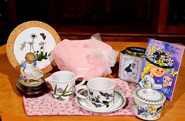 A birthday tea tableau in honor of a friend. There is Portmeirion Botanic Garden china, a bisque porcelain music box statuette of a little girl reading a book, a frilly pink cozy cover and pink calico tea towel, and a birthday card from the 1950s. The tea is Alice In Wonderland tea from Harney and Sons.