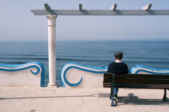 Picture of a person, seen from behind, sitting on a bench while looking at the sea.