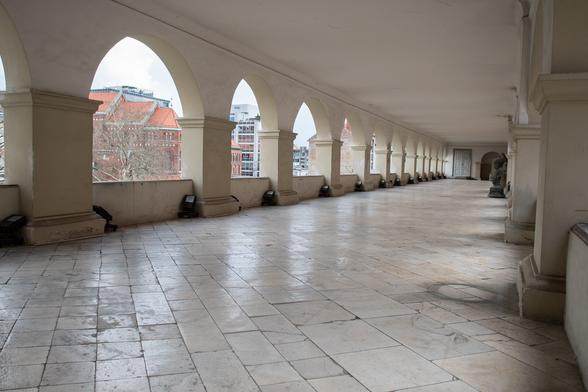 Blick in den riesigen Arkadengang im Obergeschoß von Schloss Maribor (Mariborski Grad), erkennbar sind die Rundbögen und etwa 12 wuchtige quadratische Säulen, sowie der weiße Boden, bestehend aus schräg zur Länge verlaufenden glatten Steinplatten. Ganz im Hintergrund sieht man eine graue Tür, daneben einen weiteren Rundbogen. Rechts im Vordergrund sind teilweise die ersten beiden Säulen der rechten Arkadenbögen sichtbar.

View into the huge arcade on the upper floor of Maribor Castle (Mariborski Grad), recognizable are the round arches and about 12 massive square columns, as well as the white floor, consisting of smooth stone slabs running diagonally to the length. A gray door can be seen in the far background, with another round arch next to it. On the right in the foreground, the first two columns of the right-hand arcade arches are partially visible.