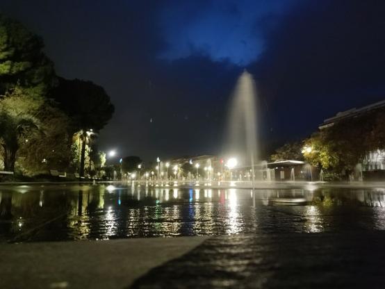 A low-angle shot of a fountain plaza in Nice. Foreground is the wet paving, framed by trees on the left and trees and buildings on the right. The background shows low streetlamps along another path through the plaza. In front of the lights are low waterspots coming out of the plaza and mid-ground is a higher waterspout.