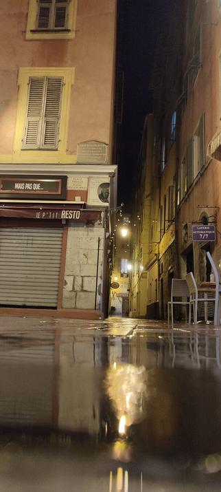A low angle of Nice old-town on a rainy night. Foreground is a puddle, left of frame is a closed restaurant, right a long building framing a narrow alleyway with hanging lights and chairs outside another closed resturant