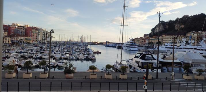A picture of Nice marina in the evening before sunset. Many tightly packed small boats on the left with multi-story buildings extreme left. behind the road in the foregound are larger cruisers and more large cruisers along the right of the marina