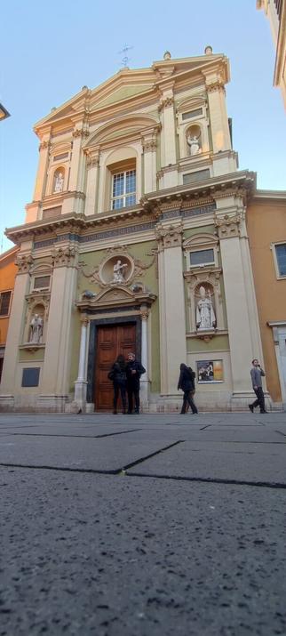 A Lw-angle view of the front of Nice cathedral. Paving low in the foreground, the wooden doors with people standing in front. Two white stone columns frame statues built into the walls either side of the door. Another statue in the wall above the ornate triangular lintle. Second story is similar with a large, leaded window above the door