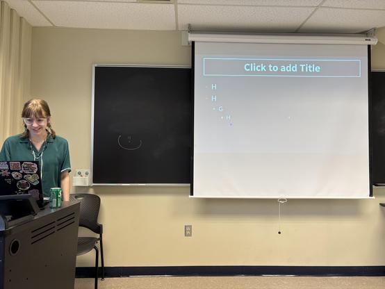 A young woman in a green top standing house left of a classroom projector screen. On the screen is a slide of a template presentation.