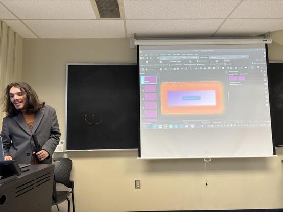 A young man in a grey jacket standing house left of a classroom projector screen. On the screen is the LibreOffice Impress program in editing mode.
