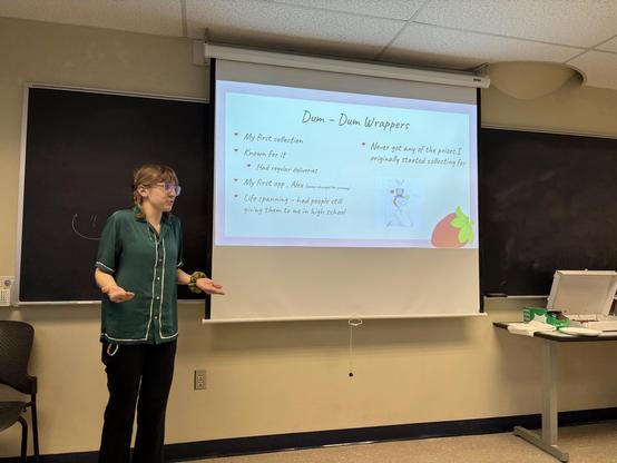 A young woman in a green top standing house left of a classroom projector screen, presenting a slide whose current section title is "Dum Dum Wrappers."