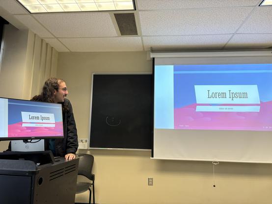 A young man in a black jacket standing house left of a classroom projector screen, with a monitor covering his torso. On the screen and on the monitor is the same image of a slide show presentation.