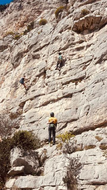 A climber looking up, midway up a limestone cliff, the belayer on a ledge at the bottom. Another climber sits on the rope to the left.