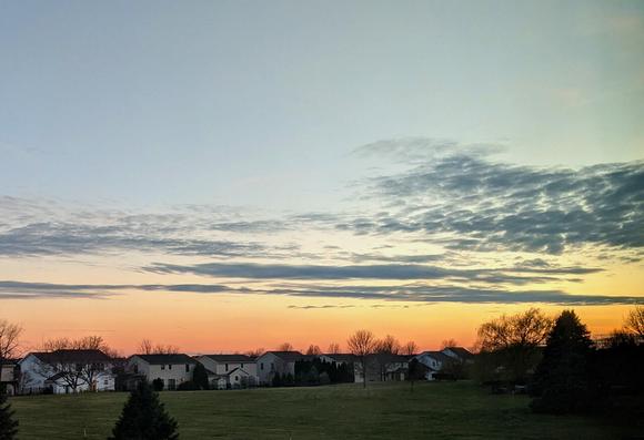 Orange sunset with pale blue sky and dark clouds over a field with trees and houses