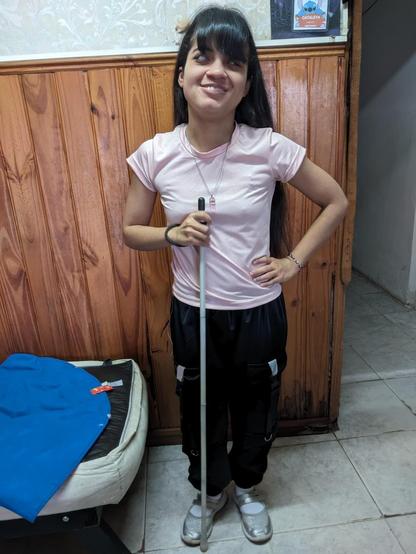 La imagen muestra a una joven de cabello largo y oscuro con flequillo, vestida con una camiseta rosa claro y pantalones negros con detalles blancos y bolsillos grandes. Ella está sonriendo y sosteniendo un bastón blanco con ambas manos, lo que indica que es una persona con discapacidad visual. Está de pie en una habitación con una pared de madera a sus espaldas, que tiene un diseño de paneles verticales. A su lado hay una silla o sofá cubierto con una tela azul. En la parte superior derecha de la imagen, hay un cartel o cuadro en la pared que parece tener un diseño colorido. El suelo es de baldosas claras, y la atmósfera general de la habitación es acogedora y sencilla.