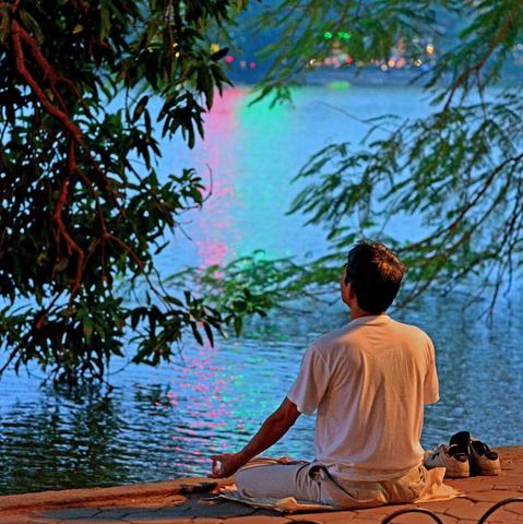Photo shows a man in white shirt sitting in meditation pose on the low wall surrounding Hoan Kiem city lake. In the background is the lake water disturbed by evening breeze, and in the background on the other side of the lake there are bokeh-typical shapes of colourful lights and buildings. The mans position is framed by low hanging green branches of trees on both sides. He sits on some sort of lightly coloured plastic mat, and his sneakers are placed neatly together to the right. It is a photo taken just after sunset, and the artificial light on the man is soft warm, while the background is a colder blue hue.