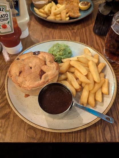 A white plate with a light brown edge. On it, there is a steak-and-ale pie on the left, mushy peas on the top, french fries on the right and a tiny sauch-pan-looking jar with a brown sauce.
on the wooden table top the is also a bottle of ketchup, mayonnaise, malt vinegar and a pint of beer.
in the background there's another plate visible, which has fish and chips on it.