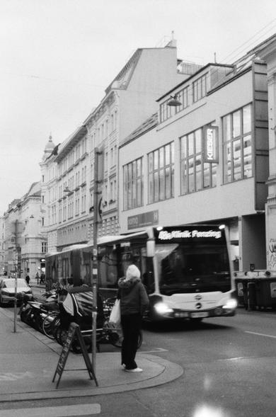 The image is a black and white street scene depicting an urban area. It features a bus moving down the street. There are also pedestrians, bicycles, and cars visible along the street.