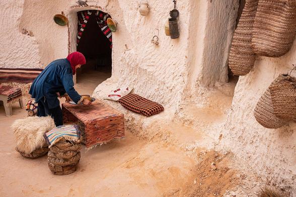 A smiling woman wearing a red headscarf and blue clothing is arranging items on a table covered with a patterned cloth in a rustic, cave-like setting, a Tunisian Berber house, with woven baskets hanging on the wall.