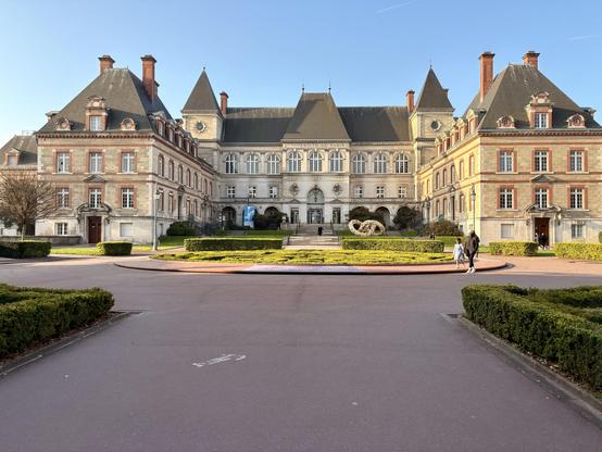 Un gigantesque bâtiment historique à l'architecture ornée avec un mélange de pierre et de brique, flanqué de jardins bien entretenus. La structure a plusieurs toits en pointe et de grandes fenêtres.