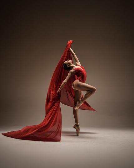 Ballerina Anna-Maria Madzharova creates a mesmerizing display of movement and grace in a studio photoshoot, wearing a striking red leotard. With a fluid ballet pose en pointe, she elegantly swings a billowing chiffon material, adding a dynamic, ethereal touch to the composition. The flowing fabric enhances the sense of motion, capturing the perfect harmony between strength, flexibility, and artistry. In this moment, dance becomes visual poetry, blending power and softness in a breathtaking expression of ballet’s beauty.