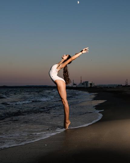 Ballerina Stefania Suciu showcases breathtaking flexibility and elegance in a sunset ballet photoshoot on the beach of Burgas during the Russian Masters Ballet summer camp. Wearing a white leotard, she moves with effortless grace, her elongated lines blending harmoniously with the golden hues of the evening sky. The soft sand beneath her feet and the rhythmic waves in the background create a serene yet powerful setting, capturing the timeless beauty of ballet in nature’s embrace.