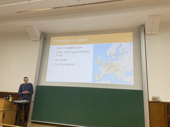 Volker Krause stellt das Projekt Transitous vor. Im Hintergrund eine Karte von Europa auf der aktuell verkehrende Züge als farbige Symbole dargestellt sind.