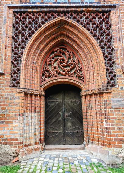 A detailed, eye-level shot showcases the ornate entrance to the brick Gothic castle chapel in Wolmirstedt, Germany. The grand Gothic arched doorway is constructed from reddish-brown bricks, characteristic of the Brick Gothic style. The deeply recessed arch features multiple brick layers, creating depth.
Above the dark, aged wooden double doors is a decorative tympanum with an intricate, swirling reddish-brown brick carving. The doors have vertical planks and large, circular metal hinges and fastenings.
Flanking the arch are brick columns. Above, decorative brickwork with a repeating quatrefoil pattern, typical of Brick Gothic architecture, forms a frieze.
The weathered reddish-brown brick facade shows visible mortar lines. The entrance is approached by a pathway of small, irregular grey and beige stones with patches of green moss. The overall impression is of historical significance and Brick Gothic architectural detail. A small, illegible sign is on the right.