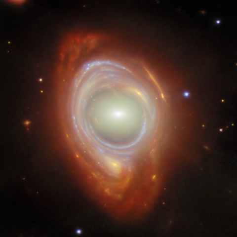In the centre is an elliptical galaxy, seen as an oval-shaped glow around a small bright core. Around this is wrapped a broad band of light, appearing like a spiral galaxy stretched and warped into a ring, with bright blue lines drawn through it where the spiral arms have been stretched into circles. A few distant objects are visible around the ring on a black background.