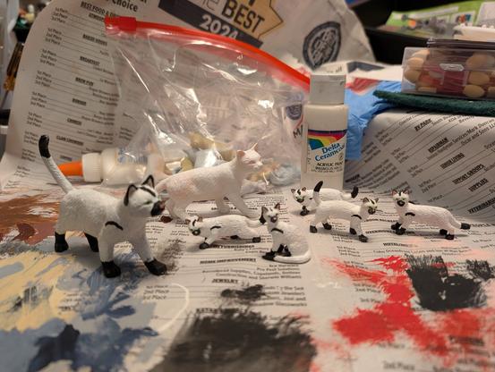 Work table covered with paint-spattered newspaper on which sit various paints and supplies, and seven plastic cats, all but one hand painted in a Minions uniform pattern. Five of these are the smaller "minions kittens."