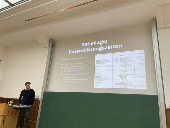 Felix Gündling präsentiert, wie barrierefreies Routing in MOTIS funktioniert. Auf Slide im Hintergrund ist eine Liste von Unterstützungszeiten an deutschen Bahnhöfen aufgeführt