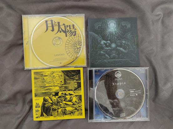 2 CDs and their covers laid out in a square.
On the left is a yellow CD, Xanvala's 1st album called "Moon and Sun" / 「月と太陽」
On the right is the dark cover of Jiluka's Xtopia.