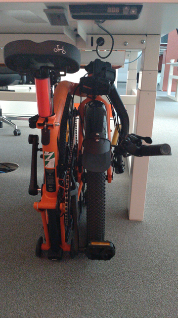 Ein zusammengefaltetes,orangenes Brompton G-Line unter einem weißen, höhenverstellbaren Schreibtisch.